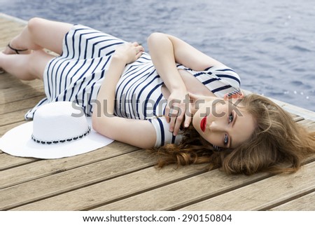 pretty young woman with fashion striped dress, hat and stylish make-up lying on wood jetty near blue water in summertime. Trendy tourist in vacation