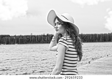 black and white shot , elegant pretty girl , in blue lines dress and summer white hat , outdoor near a lake , with some water on background