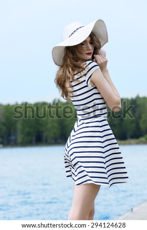 beautiful girl standing near the water on the seaside , she has elegant blue line dress and white hat . looking in camera