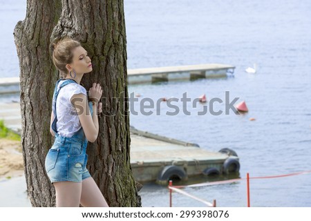 casual pretty woman with denim overalls and t-shirt posing with relaxed pose outdoor near tree with blue sea water on background