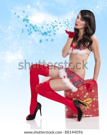 sweet christmas girl in a mini christmas outfit sitting on a present box and blowing a kiss