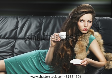 lady laying down on sofa keeping and drinking from a little cup of coffee. wearing green dress. she is lying on the sofa, looks in to the lens and takes a cup hand and saucer with left hand.