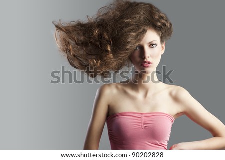 beauty fashion portrait of beautiful young brunette with curly hair flying and creative hairstyle. she looks in to the lens, has hair raised on the right and left hands on left hip.