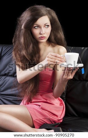 sophisticated young pretty brunette in pink elegant dress sitting on a black sofa and tasting a cup of tea. she looks at left and takes one cup of tea with both hands