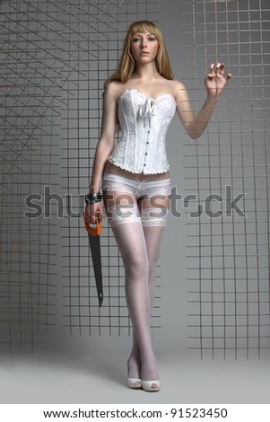Sexy cool blonde woman posing alongside re-inforcing mesh in a white corset and stockings, holding saw, studio.