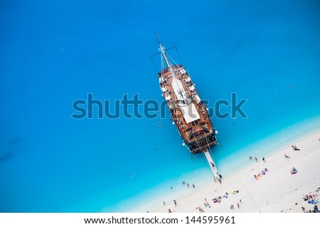 large wooden cruise sail ship anchored close to white sand beach, tourists swimming and having summer fun