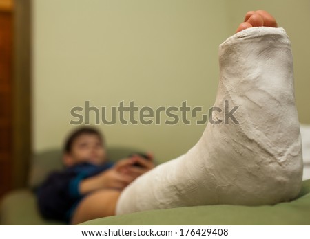 Little boy with broken leg in plaster cast lying on sofa at home and using smart phone. Focus in foreground