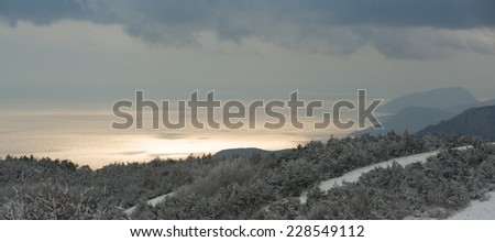Crimea Mountains covered with frosted trees are in the foreground, famous Ayu-Dag mountain is in the background. The sky is covered with clouds but sunlight is reflected in the sea.
