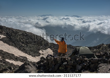 Woman dressed in orange clothes looks at cloudscape. Situation from mountain trip. Tourist is situated near the tent surrounded with stones wall.