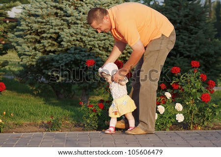 Father teaches her beautiful little daughter to walk on the background of rose bushes