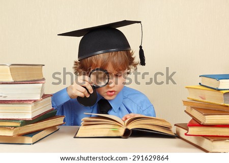 Little boy in academic hat studies an old books with a magnifier