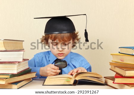 Little serious boy in academic hat studies an old books with a magnifying glass