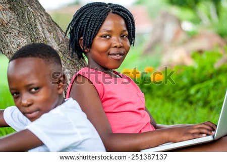 Close up portrait of cute African girl typing on laptop next to brother under tree in park.