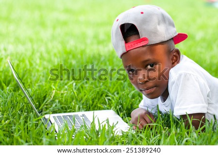 Close up portrait of African boy with baseball hat laying with laptop on grass.