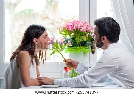Close up portrait of young man giving flower to girlfriend in restaurant. Elegant couple in white casual wear sitting next to window.