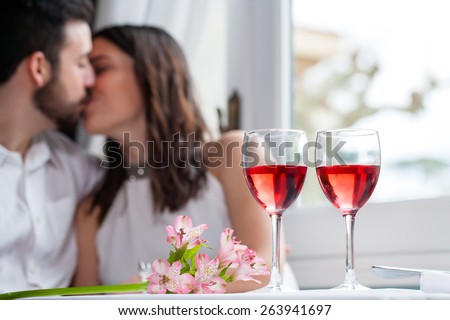 Close up detail of two wineglasses with couple in background kissing. Wine glasses filled with rose wine next to flower bouquet.