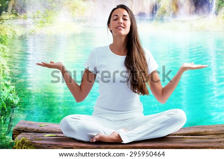 Close up portrait of attractive woman dressed in white sitting in meditating position on wooden log at blue lagoon. Young girl raising hands doing yoga.