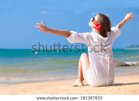 Back view young long haired woman in white shirt and sunglasses with flower in hair sitting on beach