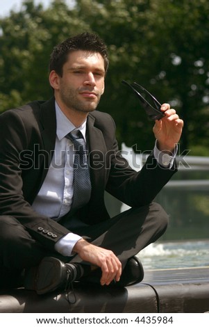 lawyer in business suit sitting cross-legged on a bench in front of a fountain taking off his sunglasses