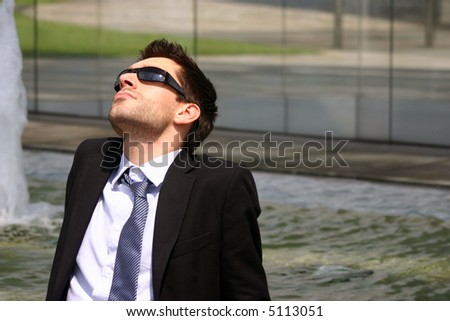 business man with designer stubble wearing sunglasses and loose necktie relaxes during his break in front of a fountain in the park