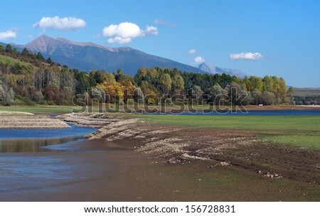 Autumn view of shore with low water level at Liptovska Mara lake in foreground. Colorful trees and Rohace mountains can be seen in the background.