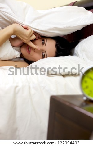 Gorgeous woman hiding herself in bed under pillow making silence sign with finger