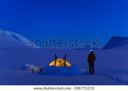 Man beside his tent in the snow looking at the sky during a cold night in Lapland (Sweden).
