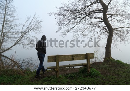 Melancholy emotions concept: sad man approaching a bench at a lake on a foggy day.