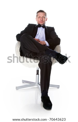 A man in a tuxedo bow tie sitting in a white chair on a black background