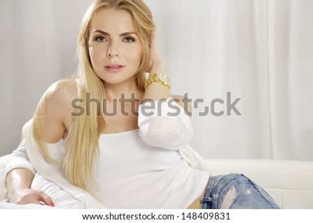 Beautiful and attractive young adult smile blonde woman posing in blue jeans and white shirt sweater on the white sofa