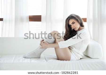 Beautiful and sexy young adult sensuality brunette woman in white sweater and grey socks on the white sofa in window on luxury apartment