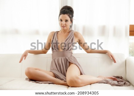 Beautiful and sexy young adult brunette woman wearing elegance fashionable dress in white sofa