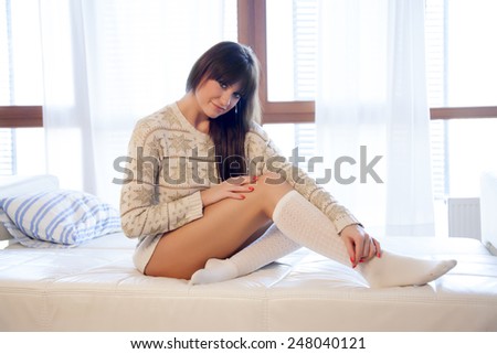 Beautiful and sexy young adult sensuality brunette woman in white sweater and socks on the white sofa in window on luxury apartment