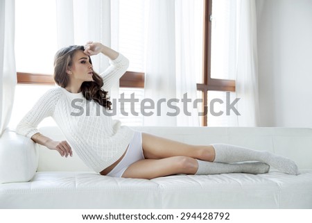 Portrait of a beautiful and sexy young adult sensuality attractive pretty brunette woman in white sweater and grey socks on the white couch in sunlight window on luxury apartment