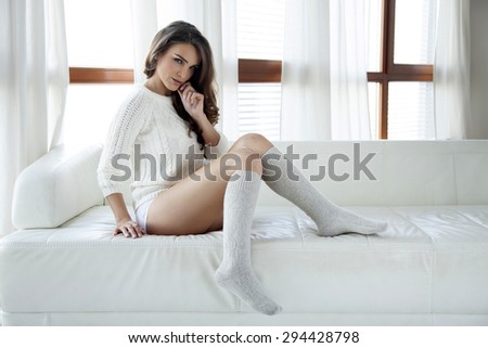 Portrait of a beautiful and sexy young adult sensuality attractive pretty brunette woman in white sweater and grey socks on the white couch in sunlight window on luxury apartment