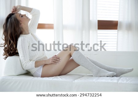 Beautiful attractive and sexy young adult sensuality pretty brunette woman in white sweater and grey socks on the white couch in sunlight window on luxury apartment
