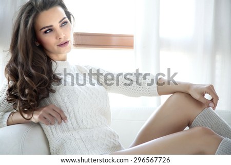 Portrait of a beautiful attractive sensuality and sexy young adult pretty brunette woman in white sweater and grey socks on the couch in morning sunlight window on luxury style hotel bedroom apartment