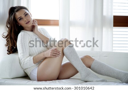Portrait of beautiful attractive and sexy young adult sensuality pretty brunette woman in white sweater and grey socks on the white couch in morning sunlight window on luxury apartment