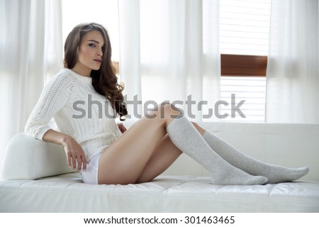 Portrait of a beautiful and sexy young adult attractive sensuality pretty brunette woman in white sweater and grey socks on the couch in sunlight window on luxury apartment