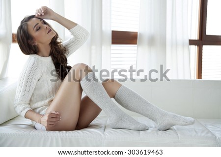 Portrait of a beautiful attractive and sexy young adult sensuality pretty brunette woman in white sweater and grey socks on the couch in morning sunlight window on luxury apartment