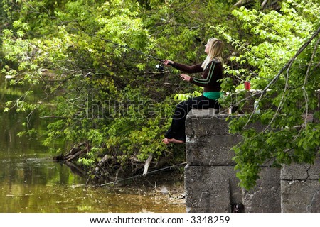 Pretty blond, barefoot woman fishing while sitting on a concrete ledge on the bank of a river.