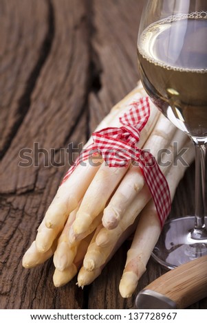 A bundle of white asparagus is on a wooden board next to a knife with a wooden handle and a glass of wine