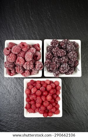 Three small porcelain dishes with frozen raspberries, currants and blackberries on a slate platter