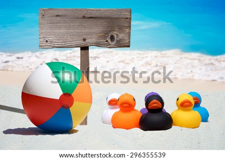Six colorful rubber ducks, beach ball and a wooden sign on the beach with the ocean in the background