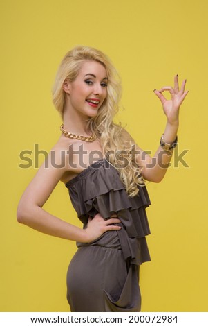 Emotional attractive young blonde woman standing in half length in brown pantsuit showing ok, isolated on yellow background