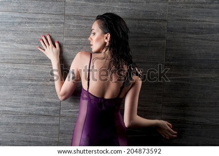Half-length portrait of very sexy dark-haired girl in great lilac lingerie standing back to us and leaning to the grey wall