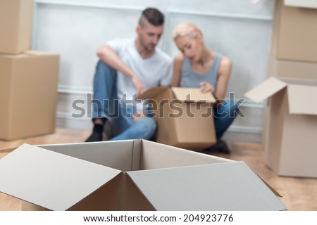 Selected focus on the big cardboard empty box lying on the floor. Tired lovely couple looking in the box on background