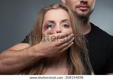 Pitiable scared young woman having black eye tearing herself away from her estranged husband standing behind her in black shirt. Isolated on grey background