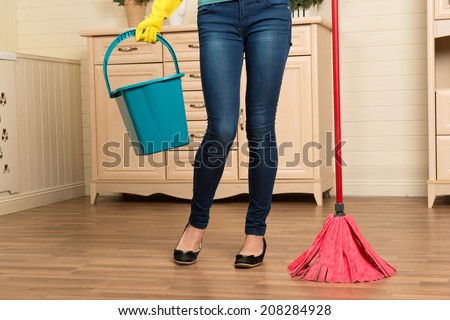 Half-length portrait of maid wearing jeans and yellow rubber gloves standing in the room and holding blue pail in one hand and mop in another