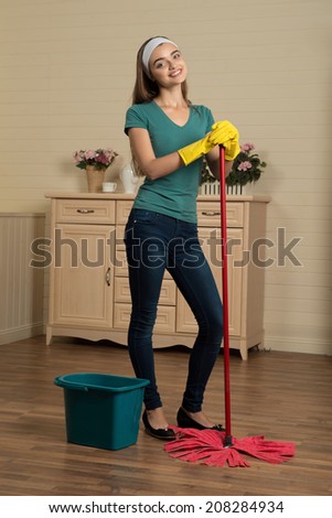 Full-length portrait of beautiful smiling housemaid wearing blue shirt and jeans standing in the room near the mop and pail